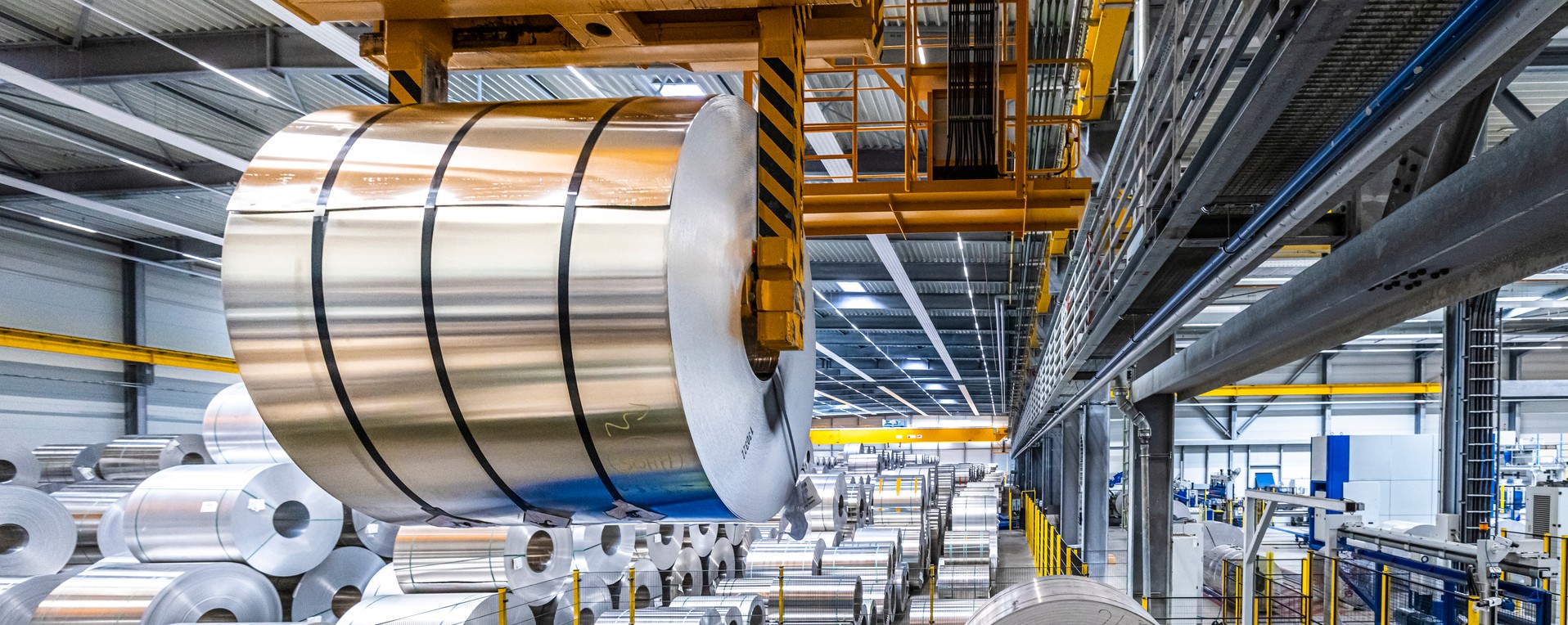 Eine großer Coil hängt an einem Kran in einer Industriehalle, umgeben von zahlreichen anderen Coils.