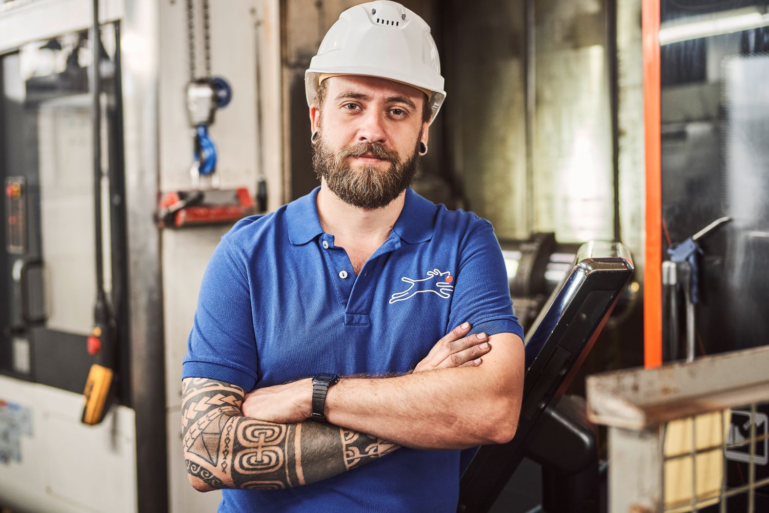 Man with beard and tattoos, wearing a white hard hat and blue polo, stands confidently with arms crossed in an industrial setting.