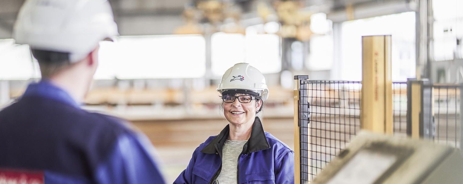 Two workers in safety gear and helmets converse in an industrial setting, with blurred machinery in the background.